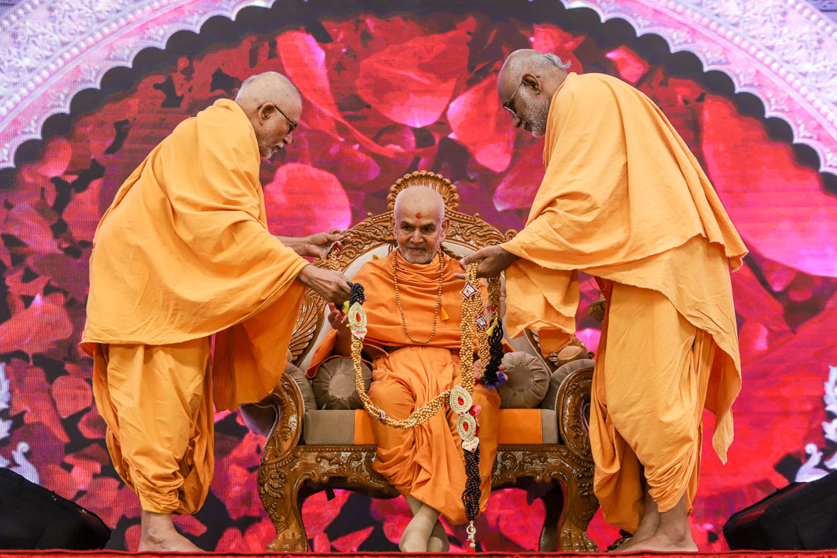 Pujya Kothari Swami and Abhayswarup Swami honor Swamishri with a garland