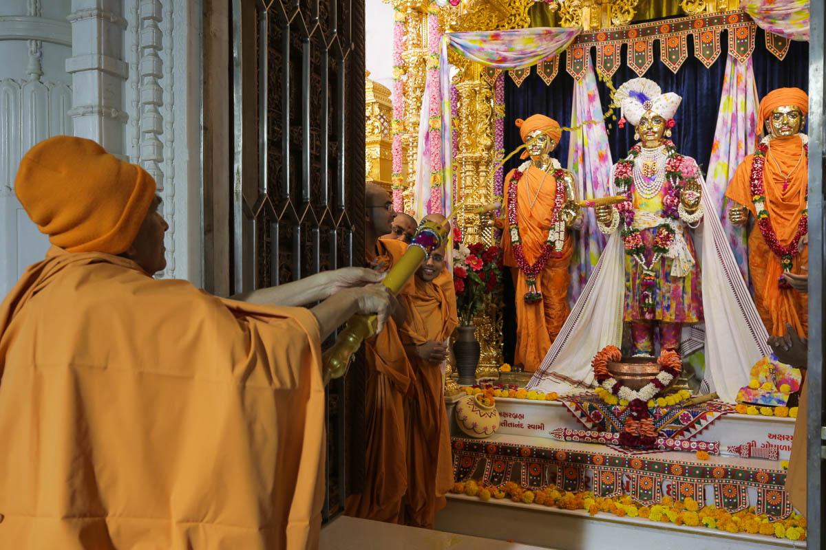Swamishri sprays saffron-scented water on Bhagwan Swaminarayan, Aksharbrahman Gunatitanand Swami and Shri Gopalanand Swami