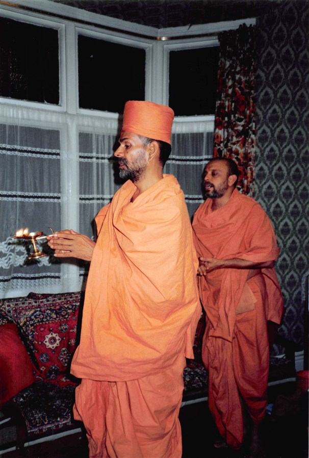 Mahant Swami and Ishwarcharan Swami perform the arti at a devotee’s home in Birmingham in 1986