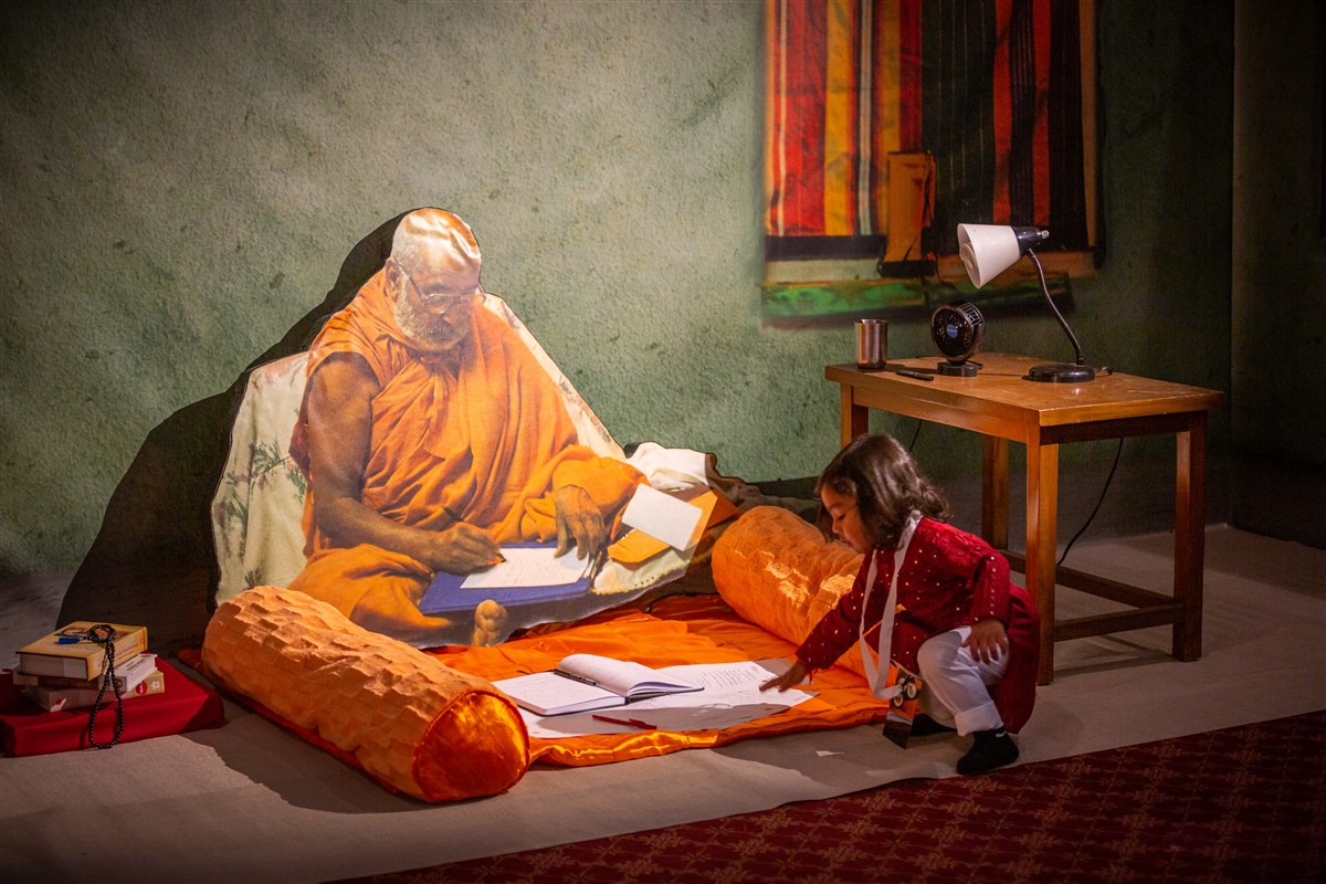 A young child reaches towards Pramukh Swami Maharaj in a heartwarming diorama of him writing one of his hundreds of thousands of letters to those in need.