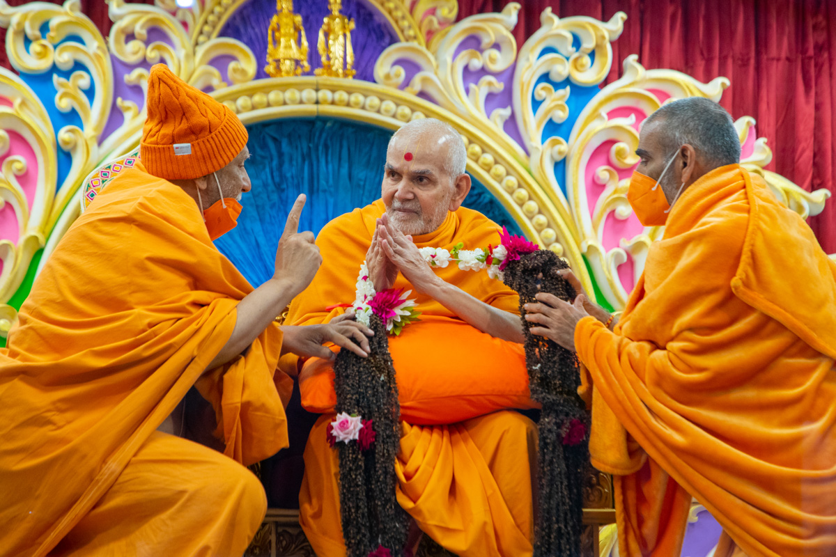 Atmaswarup Swami and Anandswarup Swami honor Swamishri with a garland