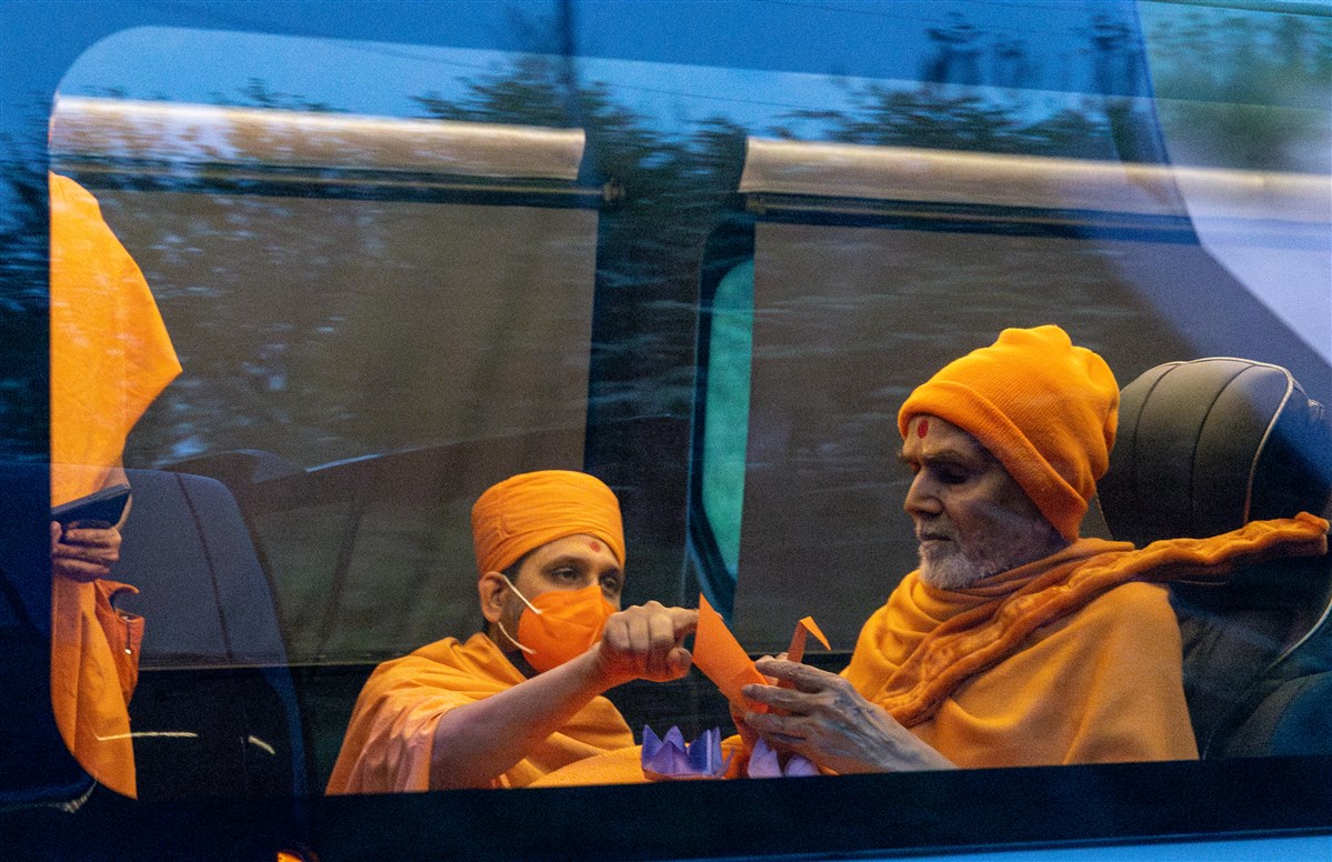 During the journey to London Mandir, Swamishri observes an origami swan and other shapes as he is informed of their symbolism during the <a href="https://www.baps.org/News/2023/Mahant-Swami-Maharaj-Joyously-Welcomed-at-BAPS-Shri-Swaminarayan-Mandir-London-23497.aspx" target="blank" style="text-decoration:underline; color:blue;">devotional welcome</a> awaiting him at London Mandir