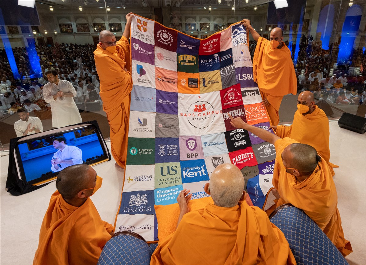Swamis present to Swamishri a decorative shawl with logos of universities attended by kishores and kishoris
