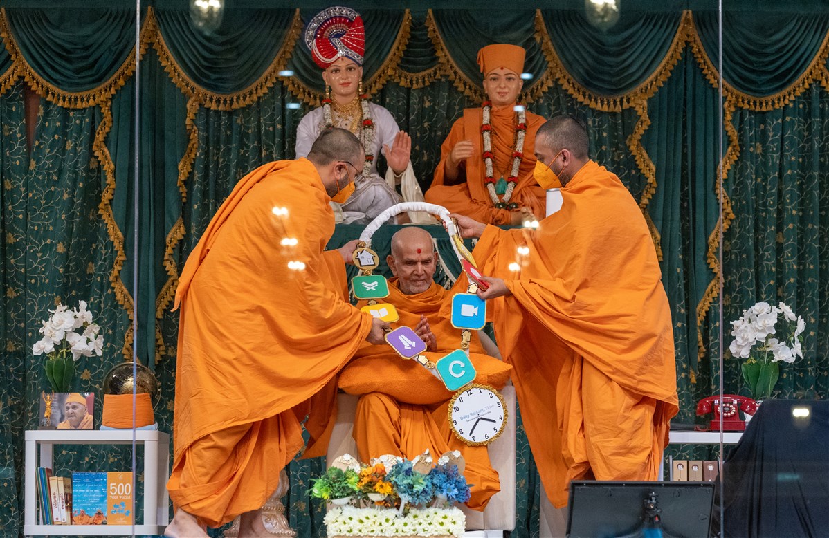 Yogikirtandas Swami and Gunkirtandas Swami honour Swamishri with a decorative garland incorporating the seven family activities of 'Daily Satsang Time'