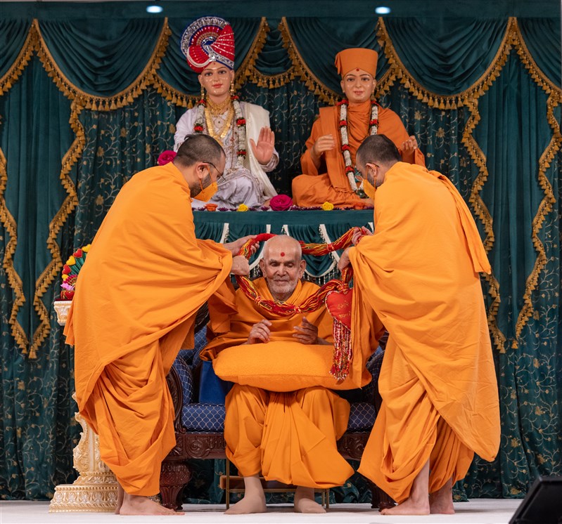 Tyagratnadas Swami and Yogikirtandas Swami honour Swamishri with a decorative garland prepared by the vadils