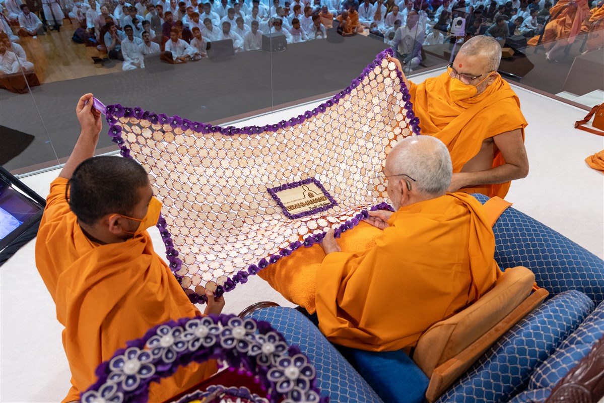 Bhadreshdas Swami and Nirdoshyogi Swami present Swamishri with a decorative shawl outlining the theme of the Yuvak-Yuvati Din celebrations, “Brahmanand: Prapti > Problems”