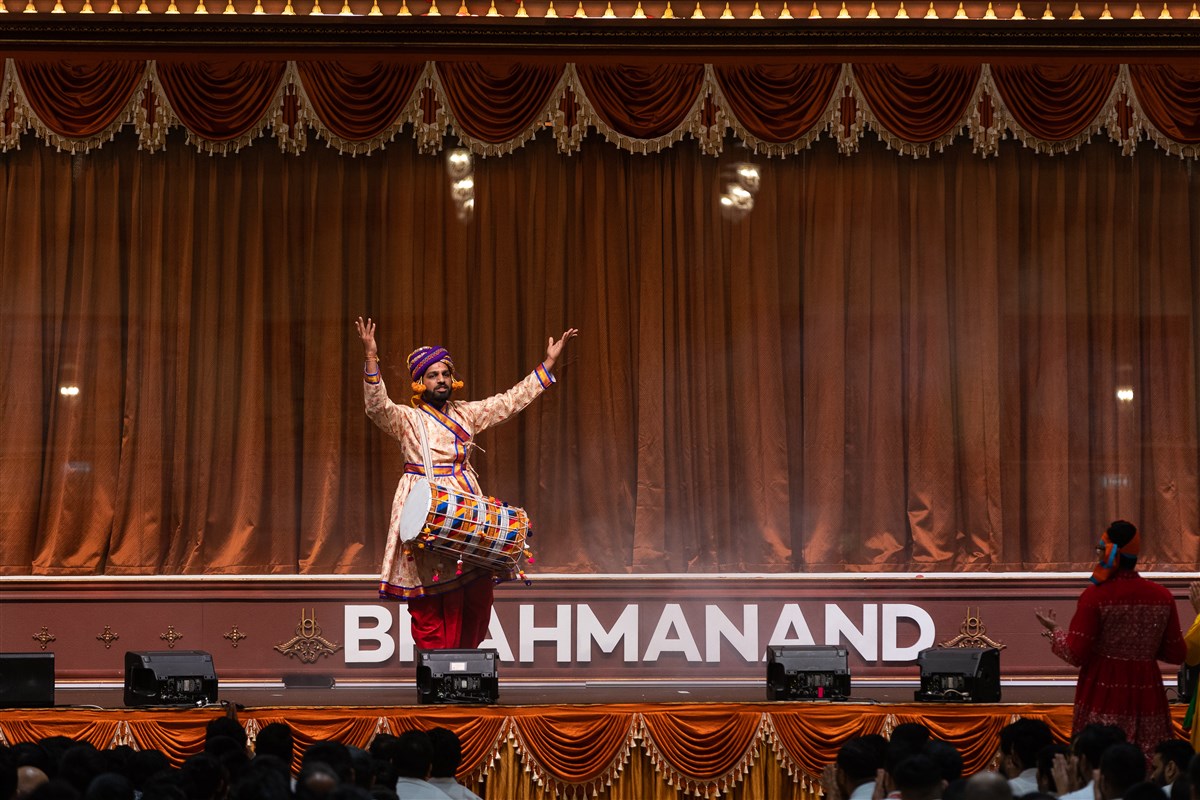 A performer announces the arrival of Swamishri in the special evening assembly