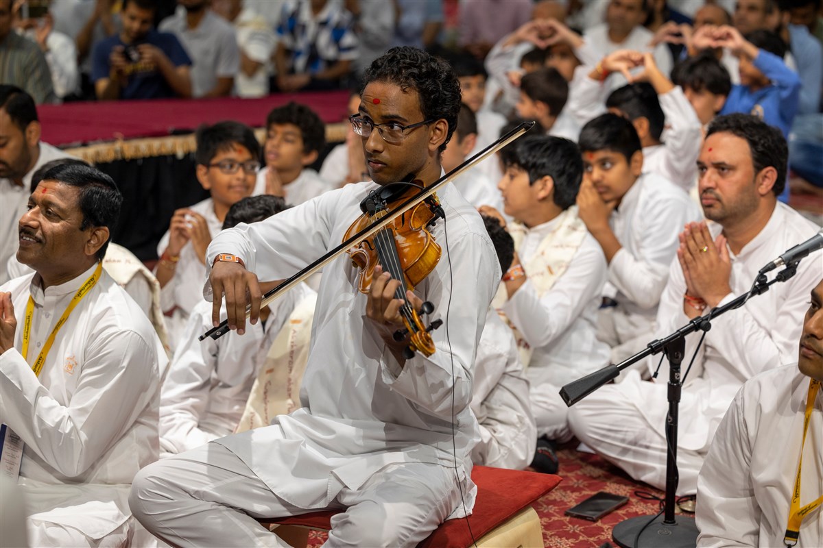 A kishore plays the violin as Swamishri greets the devotees with folded hands