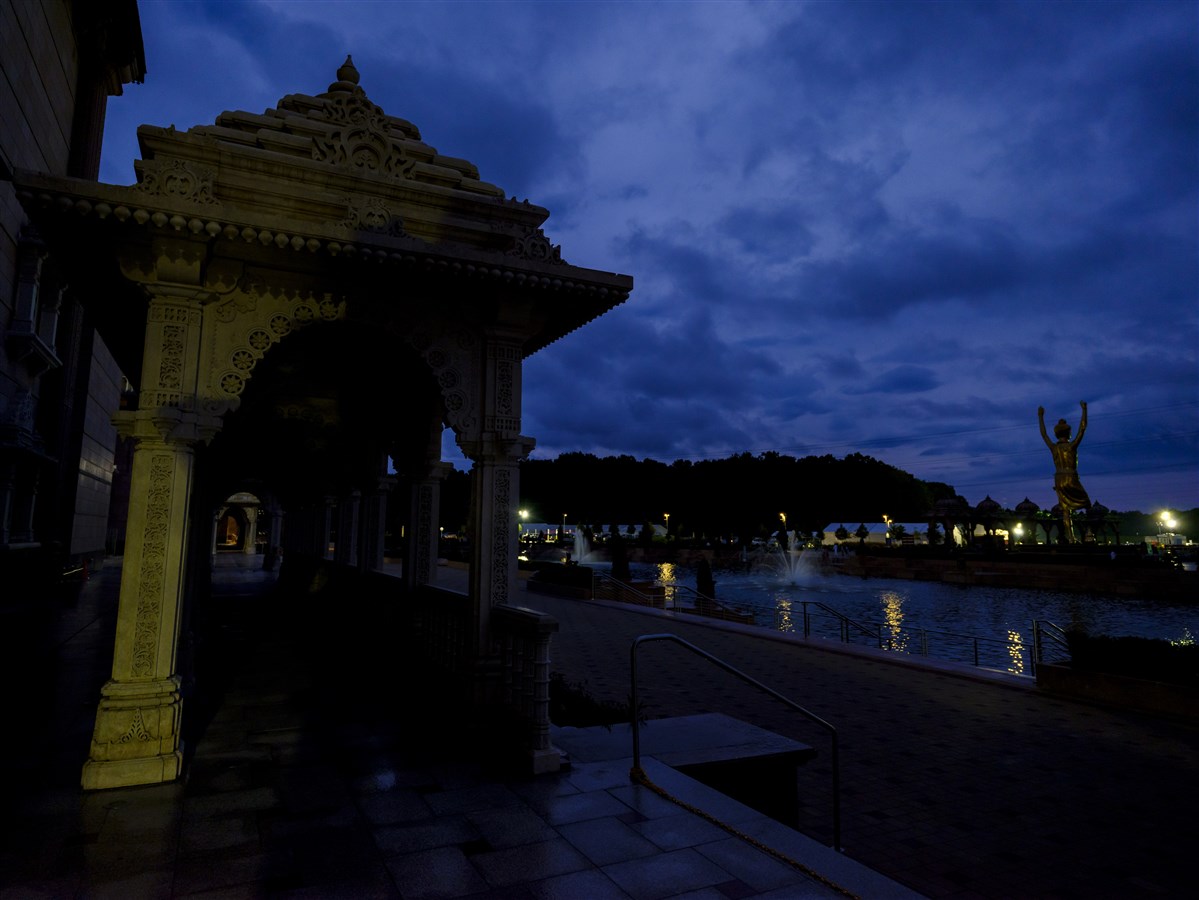 Morning hues embrace Brahma Kund as Tapomurti Shri Nilkanth Varni stands serene beneath a cloud-speckled blue sky