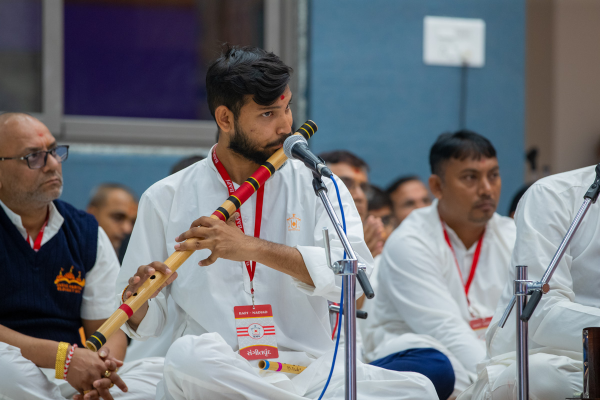 A youth plays the flute as Swamishri greets the devotees with folded hands