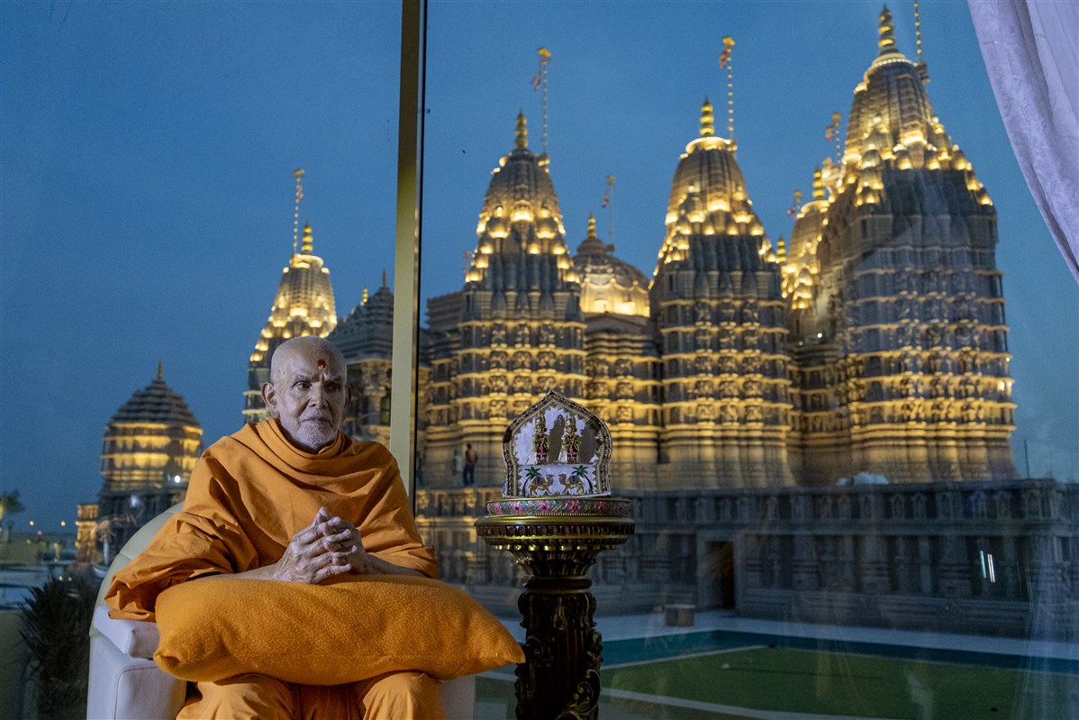 Mahant Swami Maharaj sits with folded hands, with his creation, the BAPS Hindu Mandir, in the background