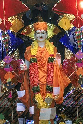 With a backdrop of kites, Shri Ghanshyam Maharaj is donned in saffron clothes of a sadhu and begging bags (jolis) in both hands